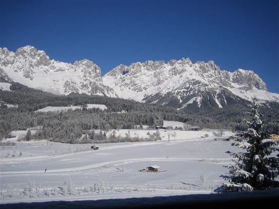 A snowy mountain landscape under a clear blue sky. In the foreground, snow-covered fields and a few small buildings can be seen.