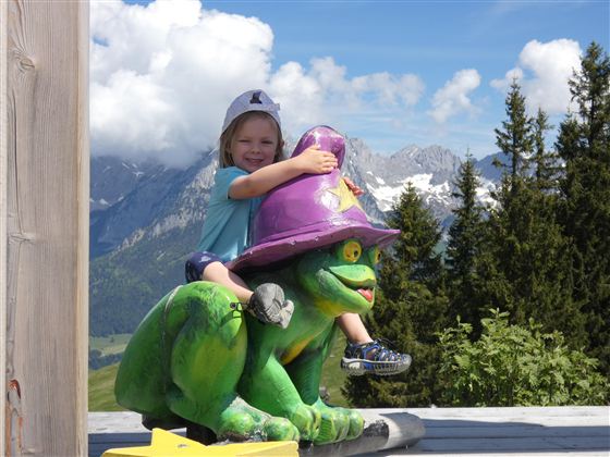 Ein kleines Kind sitzt auf einer bunten Froschfigur mit einem Hut. Im Hintergrund sind Berge und ein blauer Himmel mit Wolken zu sehen.