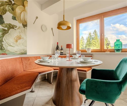 Ein einladender Essbereich mit einem runden Tisch und orangefarbenen Sitzbänken. Große Fenster bieten einen Blick auf die Natur und lassen viel Licht herein.