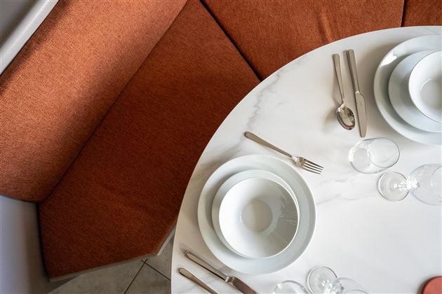 Ein eleganter Esstisch mit weißen Tellern, Besteck und Gläsern. Die orangefarbene Sitzbank bietet eine gemütliche Atmosphäre.