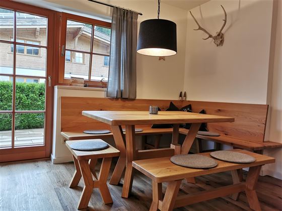 Ein gemütlicher Essbereich mit einem Holztisch und Bänken. Das Zimmer hat große Fenster, die auf eine grüne Terrasse blicken.