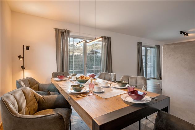 Ein modernes Esszimmer mit einem großen Holztisch und bequemen Stühlen. Große Fenster lassen viel Tageslicht herein und bieten einen Blick ins Freie.