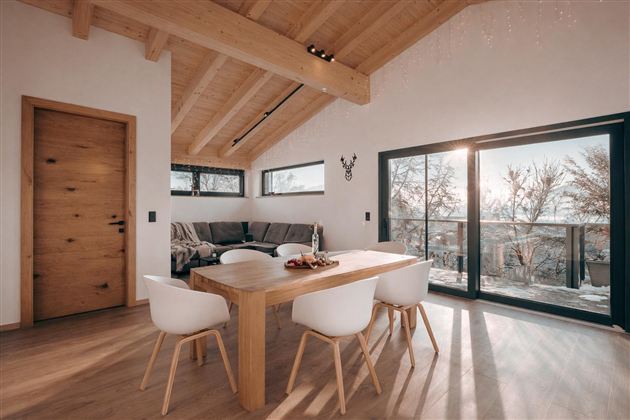Ein modernes Esszimmer mit einem Holztisch und weißen Stühlen. Große Fenster lassen Tageslicht herein und bieten einen Blick auf die Natur.