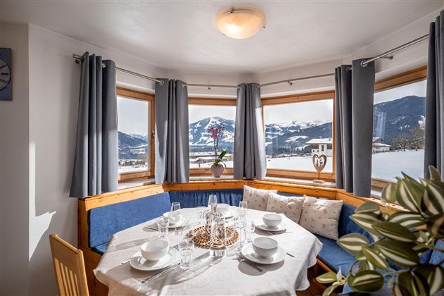 Ein gemütlicher Essbereich mit rundem Tisch und einer U-förmigen Bank. Große Fenster sorgen für eine helle Atmosphäre.