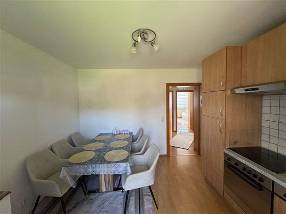 A modern kitchen with a large dining table and beige chairs. In the background, there is a door that leads to another room.