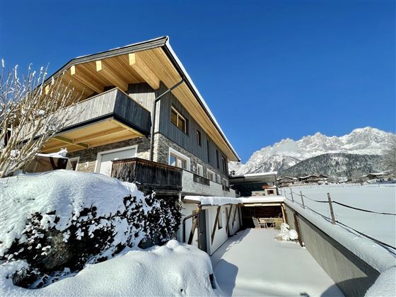 A modern house in the snow with a clear blue sky. Majestic mountains are visible in the background.