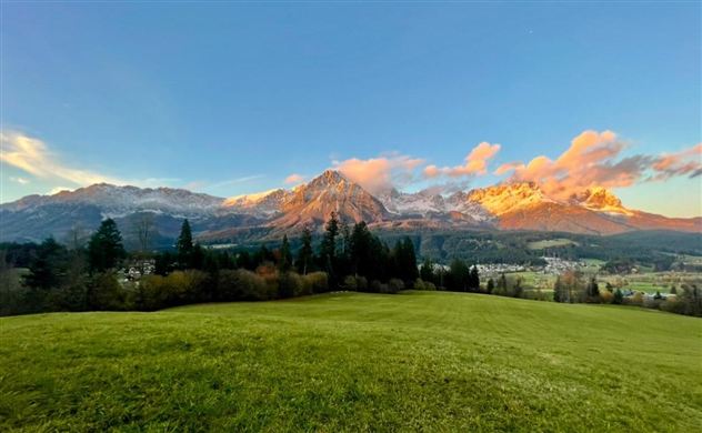 Een schilderachtig landschap met groene weiden en majestueuze bergen op de achtergrond. De lucht is helder en de wolken stralen in zachte kleuren.