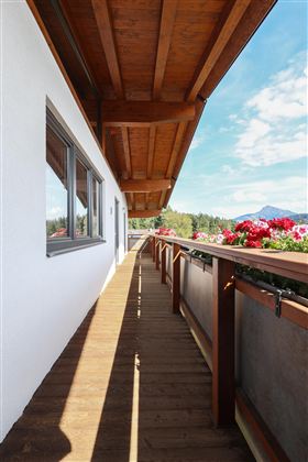 Ein heller Balkon mit Holzfußboden und Blumenkästen. Im Hintergrund sind Berge und ein blauer Himmel zu sehen.