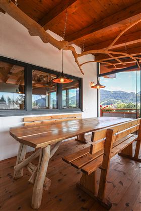 Ein gemütlicher Essbereich im Wintergarten mit einem großen Holztisch und Bänken. Die Fenster bieten einen schönen Ausblick auf die Berge.