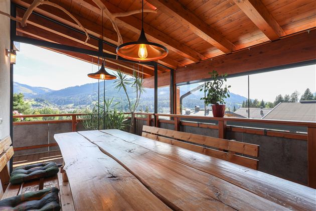Ein einladender Essbereich mit einem großen Holztisch und warmen Holzdetails. Durchfenster blickt man auf eine malerische Berglandschaft.
