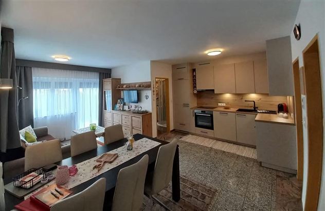 A modern living room with an open kitchen. There is a dining table with chairs and large windows that let in plenty of light.