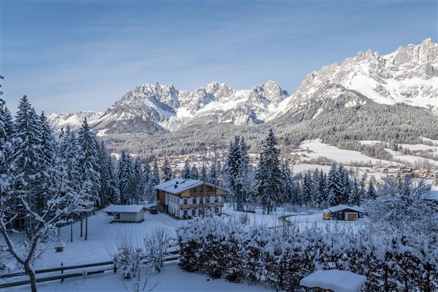 A snowy winter landscape with majestic mountains in the background. In the foreground stands a charming wooden house surrounded by snow-covered trees.