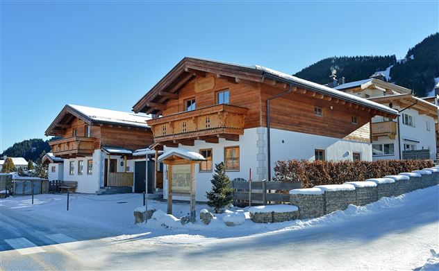 Ein schönes Chalet im Schnee, umgeben von einem klaren blauen Himmel und Bergen im Hintergrund. Die Gebäude haben traditionelle Holzelemente und eine einladende Atmosphäre.