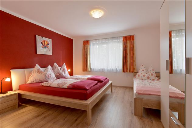 A cozy bedroom with red walls and a large bed. The room has large windows and a modern wardrobe.
