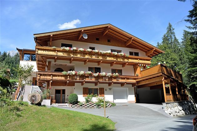 A lovely chalet in alpine style with wooden balconies adorned with carvings and beautiful flowers. Surrounded by trees and a green meadow.