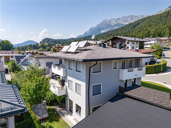 A modern house with several floors and solar panels. In the background, green mountains and a quiet residential area can be seen.