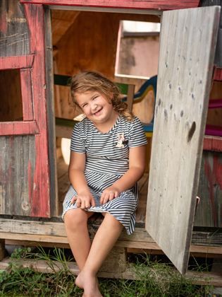 A happy girl is sitting in a small playhouse. She is wearing a striped dress and is smiling.