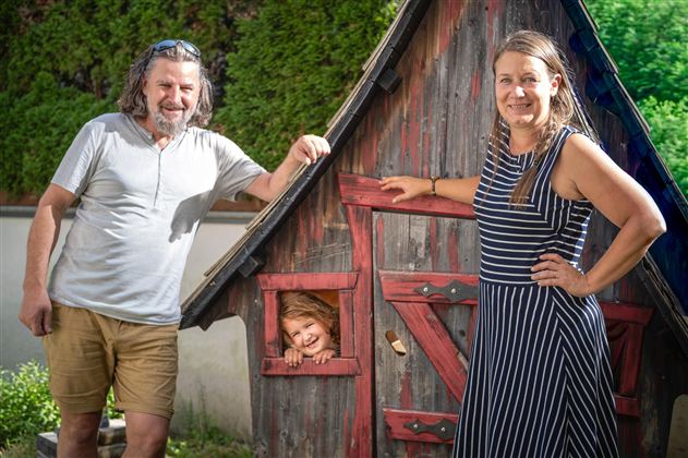 A happy couple stands next to a small wooden house. A child looks cheerfully out of the window of the house.