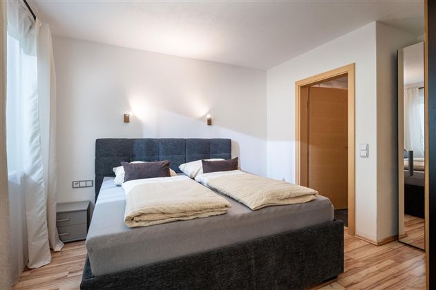 A cozy bedroom with a double bed and soft bedding. There are light walls and a large mirror that additionally brightens the room.