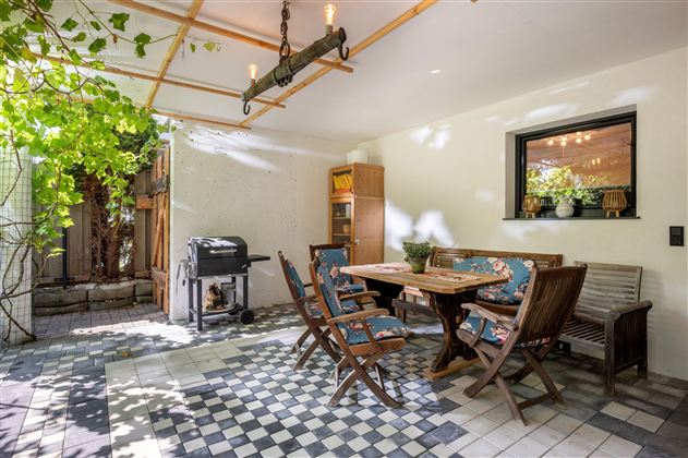 A cozy terrace with a dining table and wooden chairs. In the background, there is a grill and a plant providing shade.