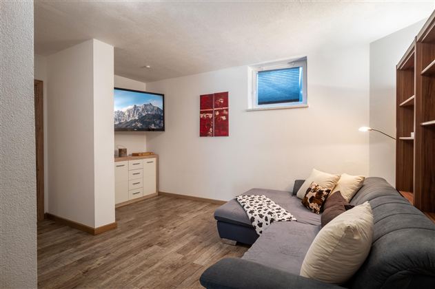 A cozy lounge with a dark blue sofa and a television on the wall. The room has wooden flooring and is brightly lit.