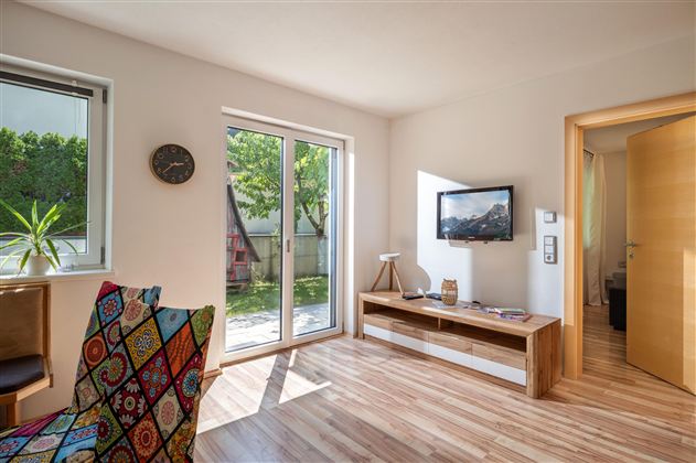A modern living room with large windows that let in a lot of daylight. There is a television, a wooden cabinet, and a colorful combination of chairs.