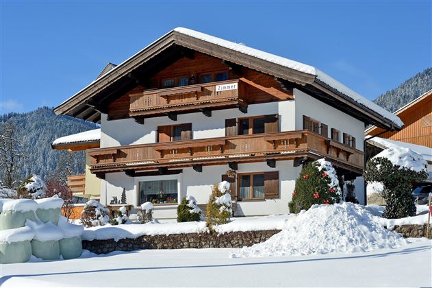 Ein wunderschönes Chalet im Schnee mit einem Holzbalon und grünen Sträuchern. Der klare blaue Himmel ergänzt die winterliche Szenerie.