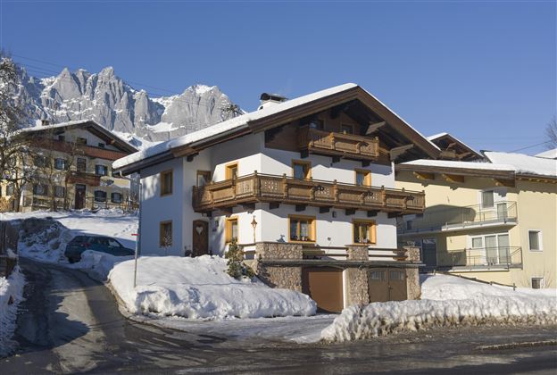 Ein charmantes, weißes Chalet mit Holzbalustraden in einer schneebedeckten Landschaft. Im Hintergrund ragen majestätische Berge in den klaren blauen Himmel.