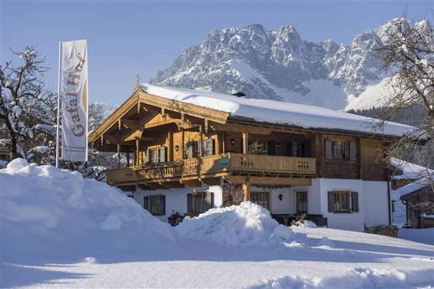 Ein traditionelles Holzhaus im Schnee, umgeben von Bergen. Die Szene zeigt eine ruhige Winterlandschaft.