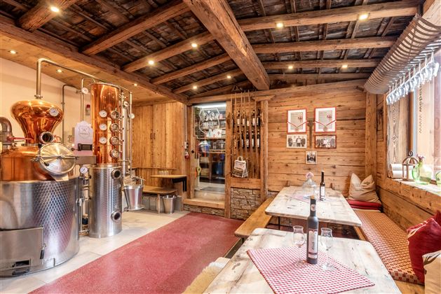 Ein gemütlicher Raum mit rustikalem Dekor und Holzwänden. Eine Brennerei steht im Vordergrund, während Tische mit roten Tischtüchern zum Sitzen einladen.