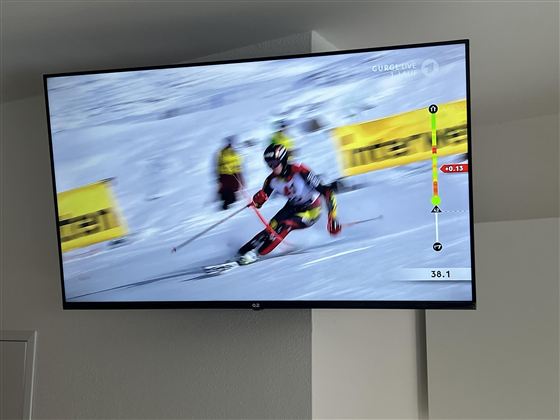 Een skiër daalt snel de piste af. Op de achtergrond zijn toeschouwers en banners zichtbaar.