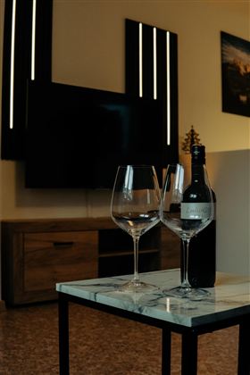 An elegant table with two wine glasses and a bottle of wine. In the background, a television and modern wall lighting can be seen.