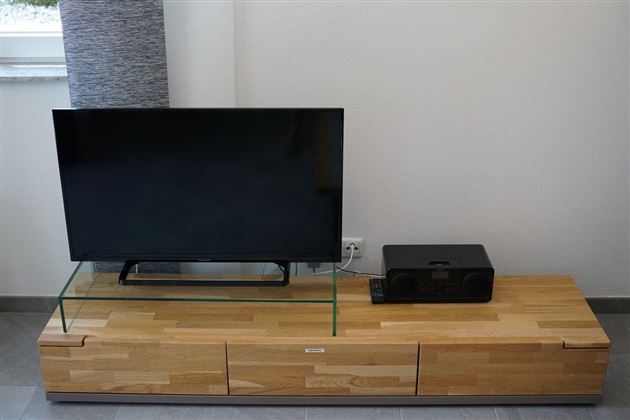 Een moderne televisie staat op een houten TV-standaard met een glazen blad. Rechts van de televisie bevindt zich een zwart geluidssysteem.