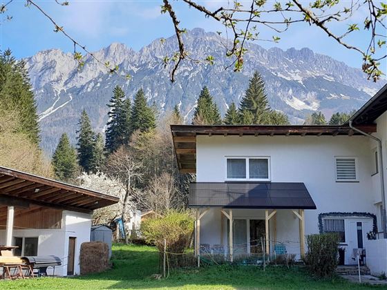 Eine ruhige Wohngegend mit einem weißen Haus und einem grünen Garten. Im Hintergrund erheben sich majestätische Berge unter einem klaren Himmel.