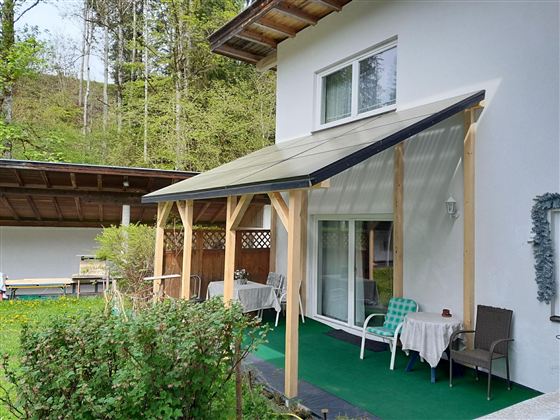 Eine überdachte Terrasse mit weißen Stühlen und Tischen. Der Hintergrund zeigt eine weiße Wand und eine Doppel-Glastür.