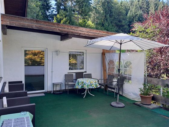 Een gezellige terras met tuinmeubels en een parasol. Op de achtergrond zijn groene bomen te zien.
