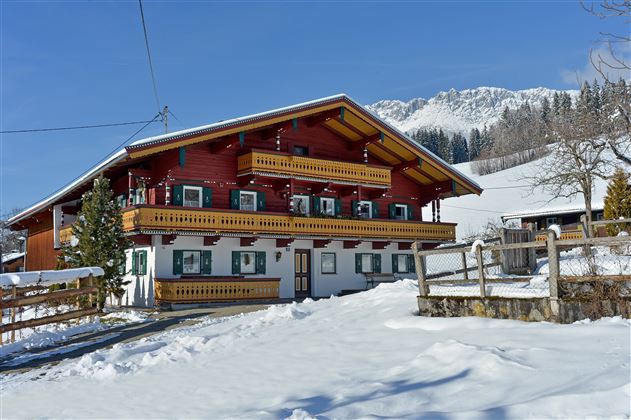 A beautiful red and white painted farmhouse in the snow. In the background, there are snow-covered mountains and a clear blue sky.