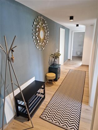 Ein moderner Flur mit einer grauen Wand und einem stilvollen Spiegel. Auf dem Boden liegt ein Teppich mit chevron-Muster und es stehen einige Möbelstücke, darunter eine Garderobe und ein kleiner Tisch.