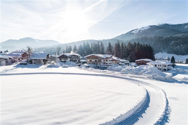 Eine verschneite Landschaft mit Häusern und Bergen im Hintergrund. Die Sonne scheint über die Winterlandschaft.