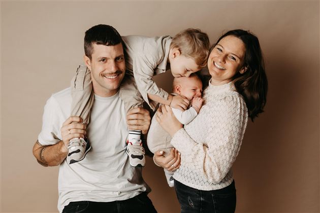 Eine fröhliche Familie posiert zusammen. Der Vater trägt ein Kind auf den Schultern, während die Mutter das Baby in ihren Armen hält.