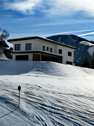 A modern house in a snowy landscape. In the background, mountains and blue sky can be seen.