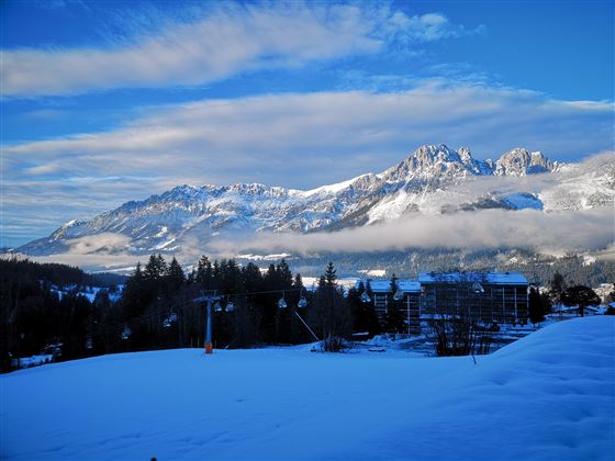 A winter landscape with snow-covered mountains and a clear blue sky. In the foreground is a snow-covered area, while clouds hang over the mountains.