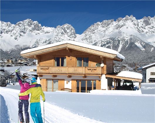 A cozy wooden house in the snow, surrounded by snow-covered mountains. Two skiers are enjoying the winter landscape.