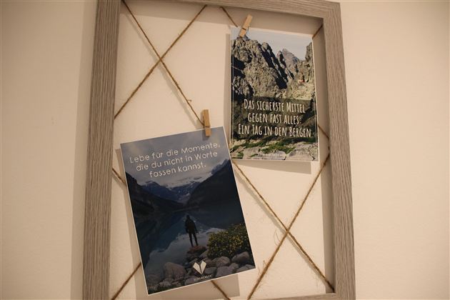 Zwei Postkarten hängen an einem Rahmen. Die linke Karte zeigt eine Person am Ufer eines Sees, während die rechte Karte einen inspirierenden Spruch in der Natur präsentiert.