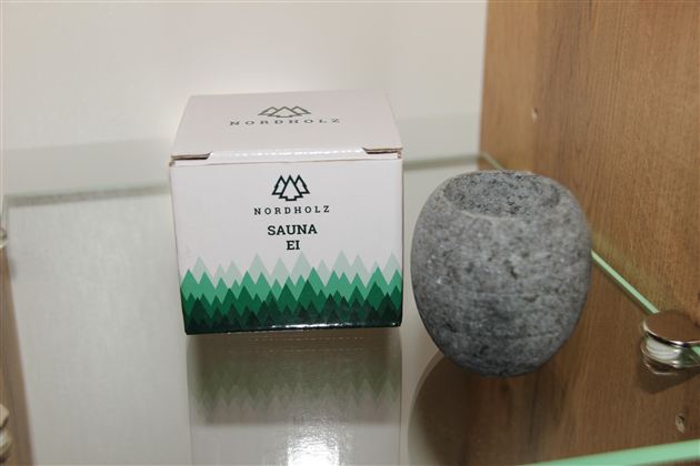 Eine Verpackung mit dem Aufdruck „Sauna Ei“ steht neben einem runden, grauen Steinobjekt. Beide Gegenstände sind auf einer glatten Oberfläche platziert.