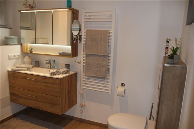 Ein modernes Badezimmer mit einem Holzschrank, einem Spiegel und Handtüchern. Es gibt eine Dusche, eine Toilette und einige dekorative Elemente.