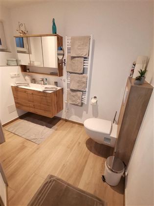 Ein modernes Badezimmer mit einem Holzwaschtisch, einem Spiegel und Handtüchern. Auf dem Boden liegt ein Teppich und die Wände sind in neutralen Tönen gehalten.