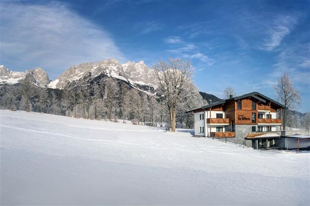 A charming house in a snow-covered landscape with majestic mountains in the background. The trees are frosty and the sky is clear and blue.