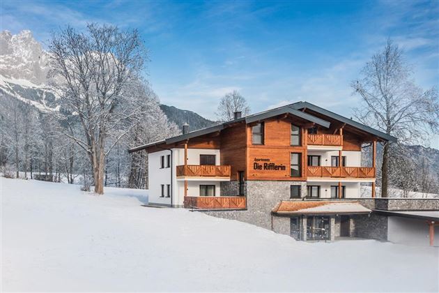 A beautiful building in the snow with a traditional wood design. The surroundings are surrounded by wintry mountains and trees.