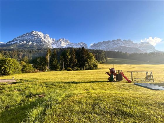 Een uitgestrekt veld met een speelplaats en indrukwekkende bergen op de achtergrond. De lucht is helder en blauw, het landschap oogt idylisch.